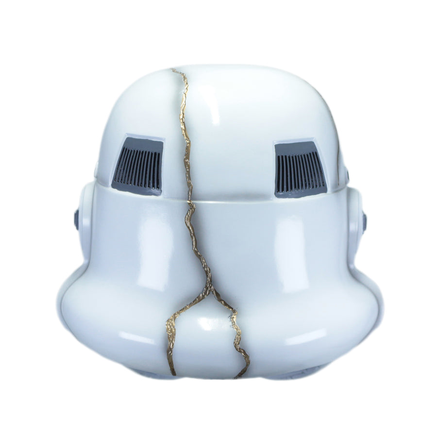 A white stormtrooper helmet with a decorative gold crack running vertically down the center and dark gray vent details.